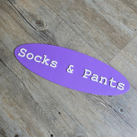 A purple, oval-shaped 3D-printed sign featuring the white, raised text "Socks & Pants," resting on a light-colored wood grain background.