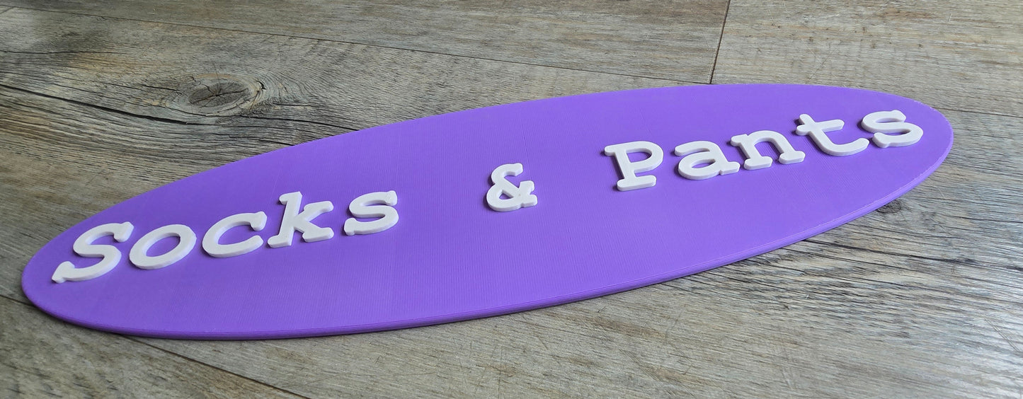 A purple, oval-shaped 3D-printed sign featuring the white, raised text "Socks & Pants," resting on a light-colored wood grain background.