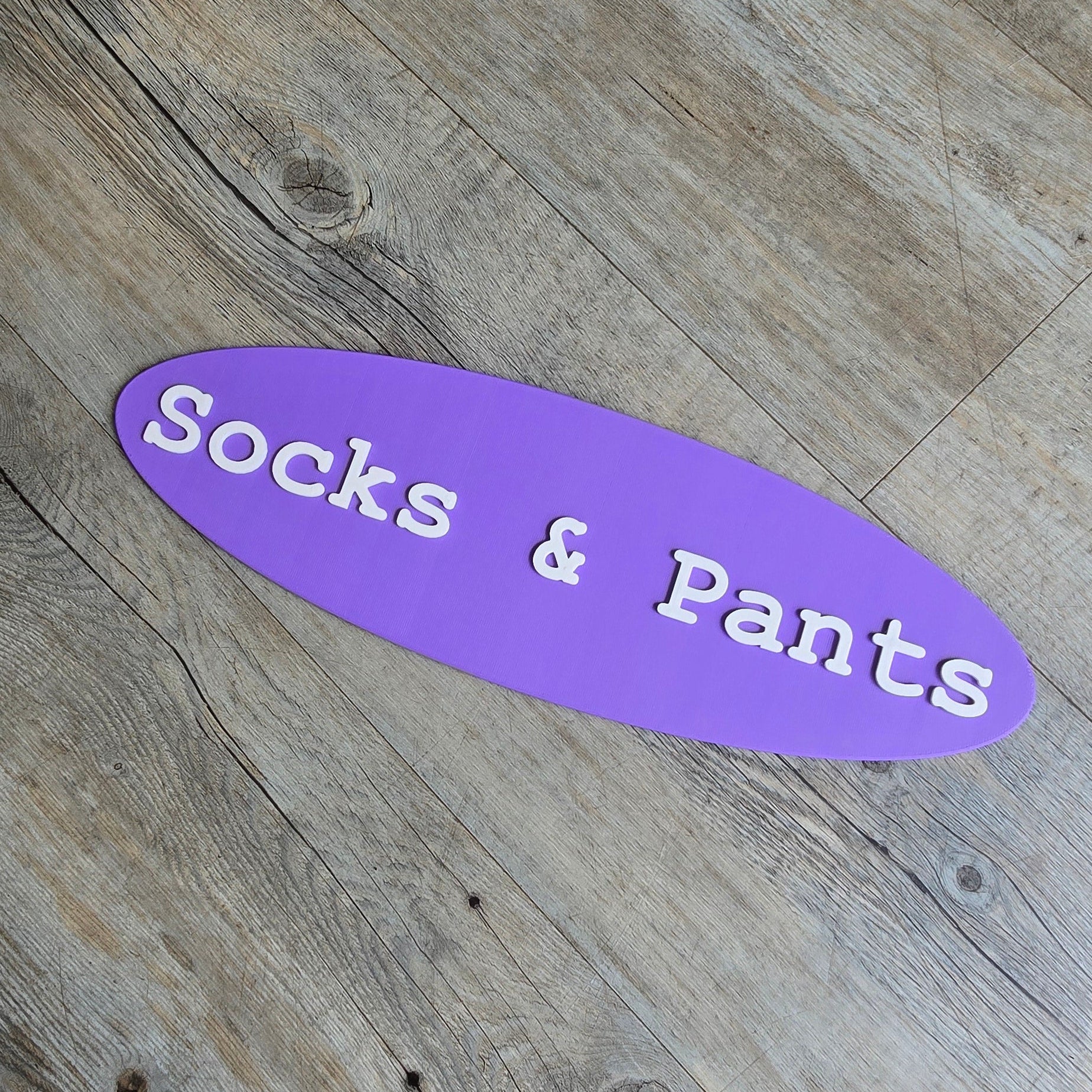 A purple, oval-shaped 3D-printed sign featuring the white, raised text "Socks & Pants," resting on a light-colored wood grain background.
