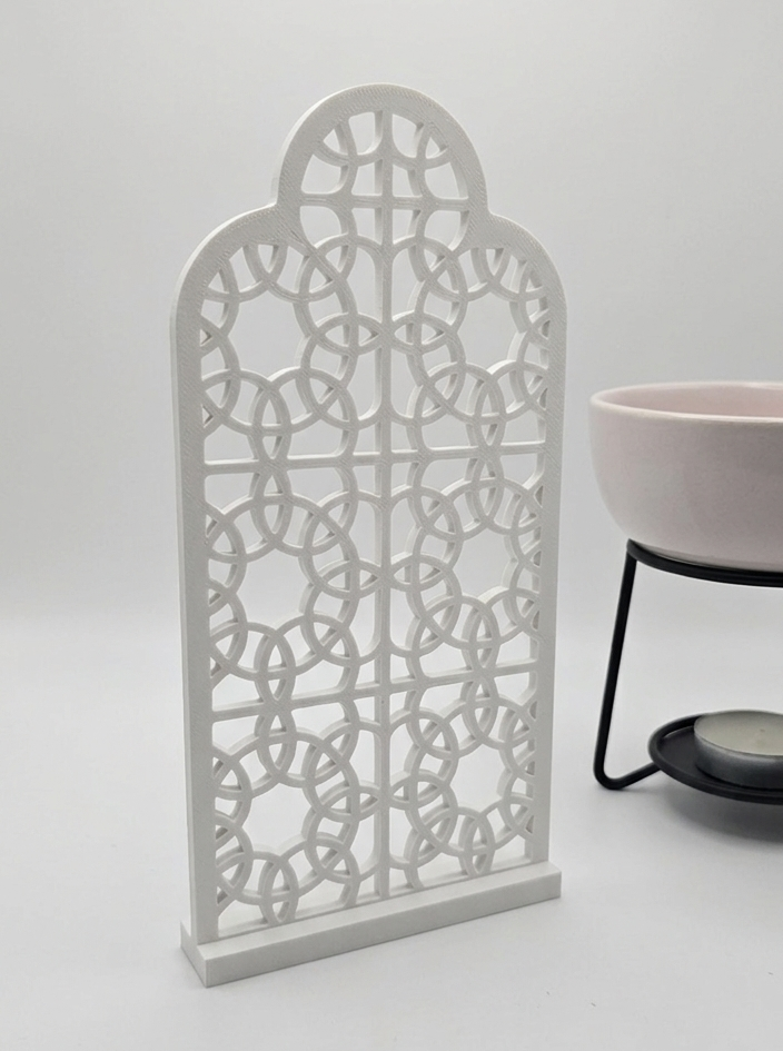 A white, 3D-printed ornamental screen with an intricate geometric arch design, standing on a plain white surface next to a light pink bowl on a metal stand.