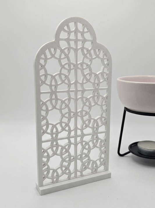 A white, 3D-printed ornamental screen with an intricate geometric arch design, standing on a plain white surface next to a light pink bowl on a metal stand.